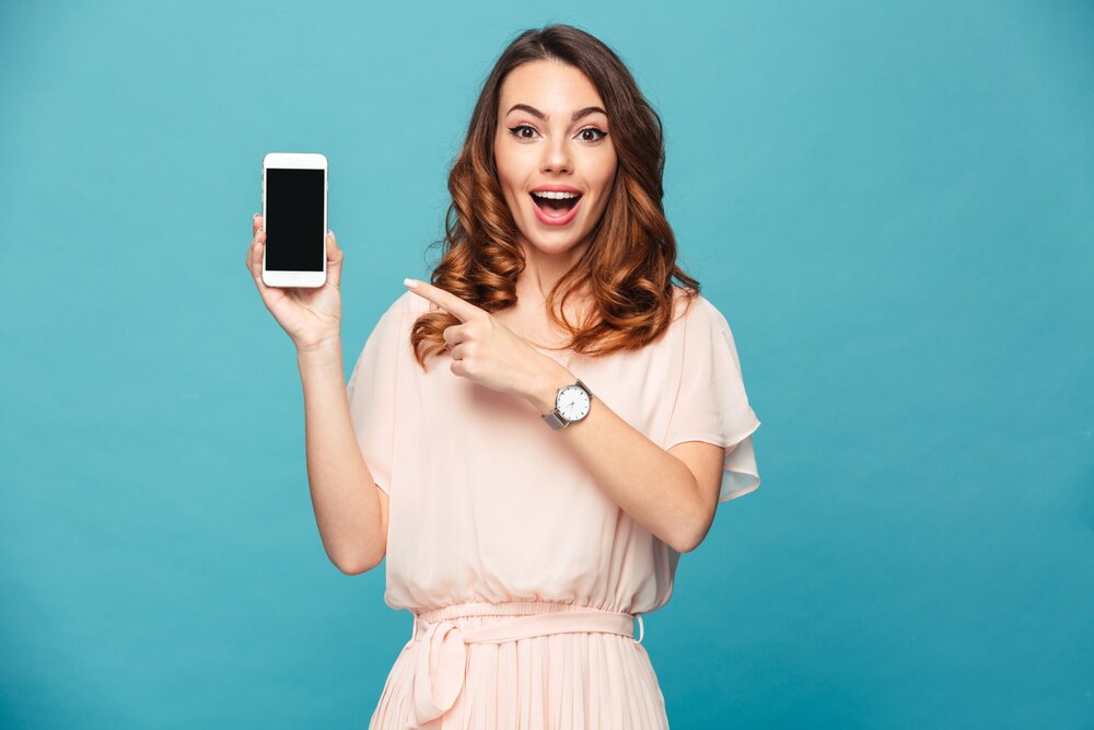 mujer mira a camara sonriente señalando smartphone